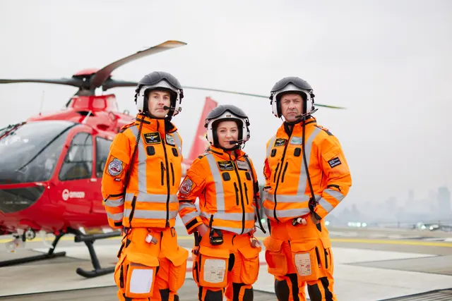 Three London Air Ambulance crew members in orange flight suits standing in front of their red helicopter