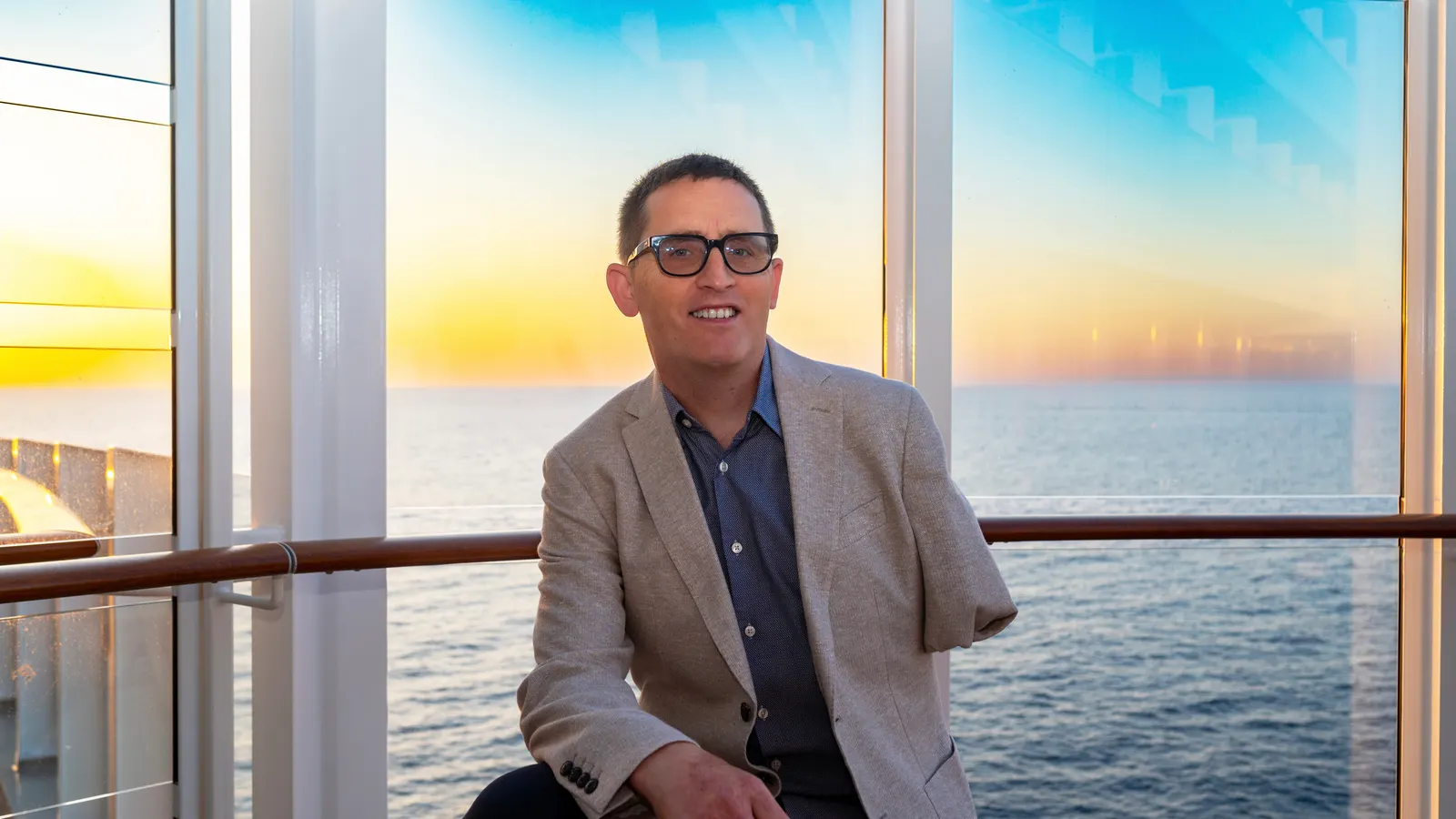 Simon Wilson smiling, sitting in front of a window with the sea and a sunset behind him