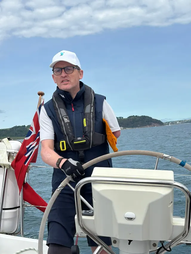 Simon at the helm of a yacht, wearing a Stride Forward cap and life jacket